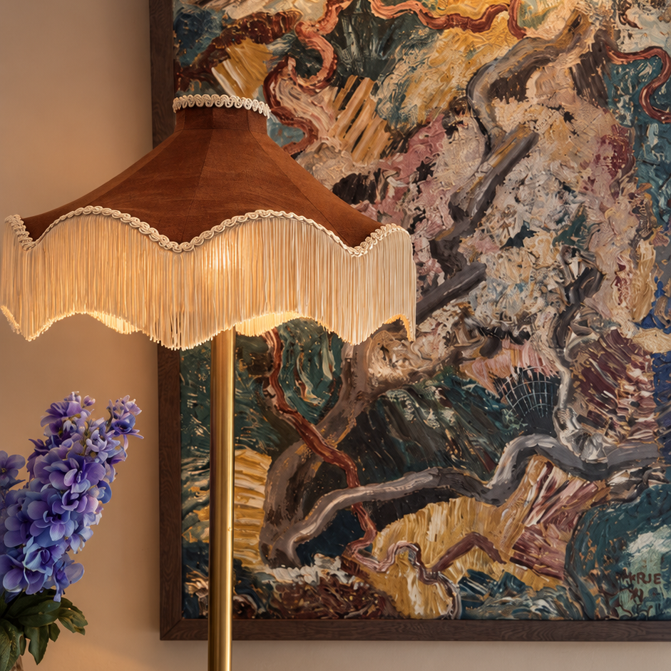 The Daisy Moiré cognac lampshade with cream fringe on a floor lamp, in front of an abstract painting and blue flowers