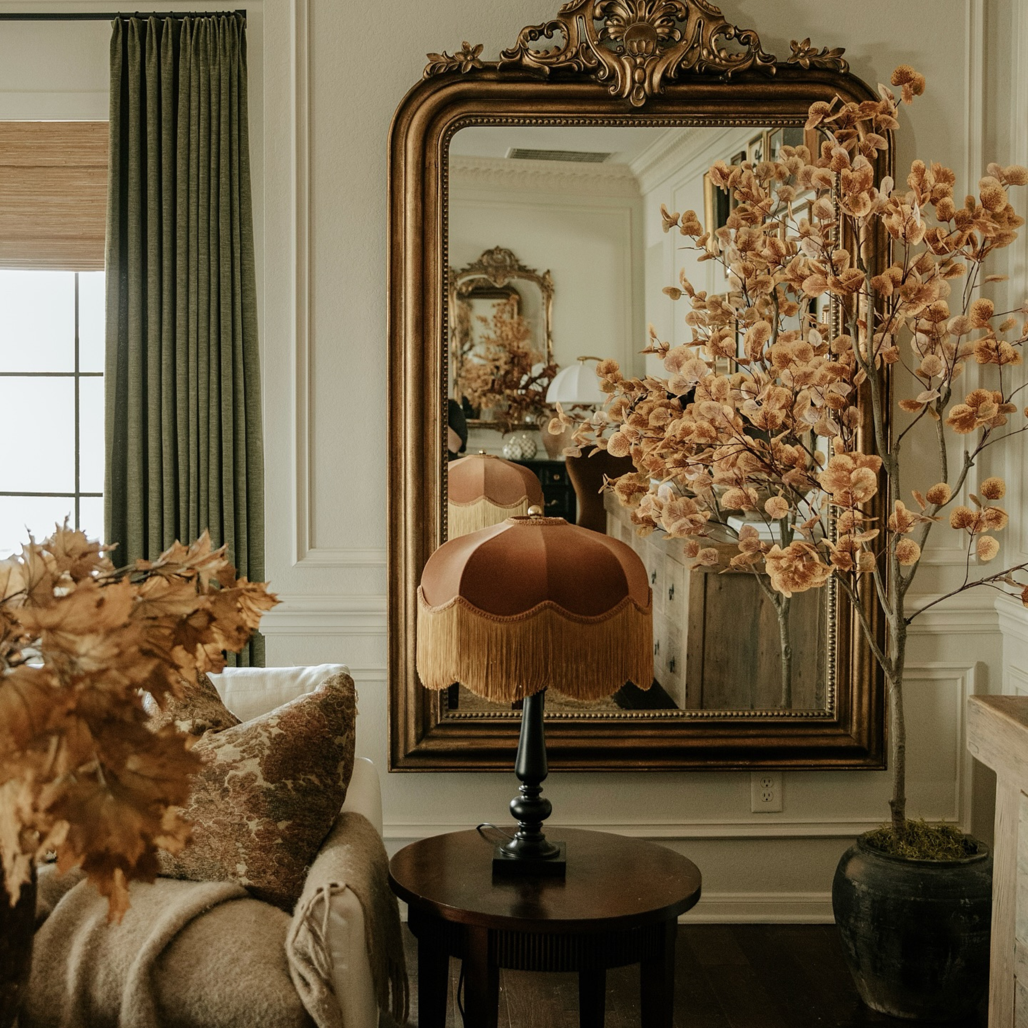 An ornate gold-framed wall mirror hangs on a white paneled wall in a styled room, with a brown fringed table lamp on a small round table below and dried floral arrangements in the foreground.