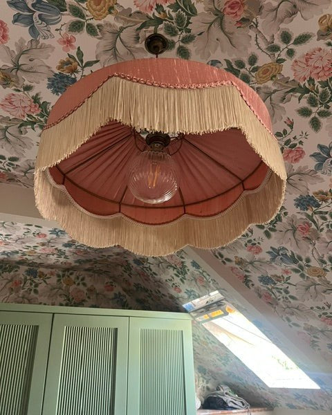 Pink fringed pendant shade under a floral wallpapered attic ceiling