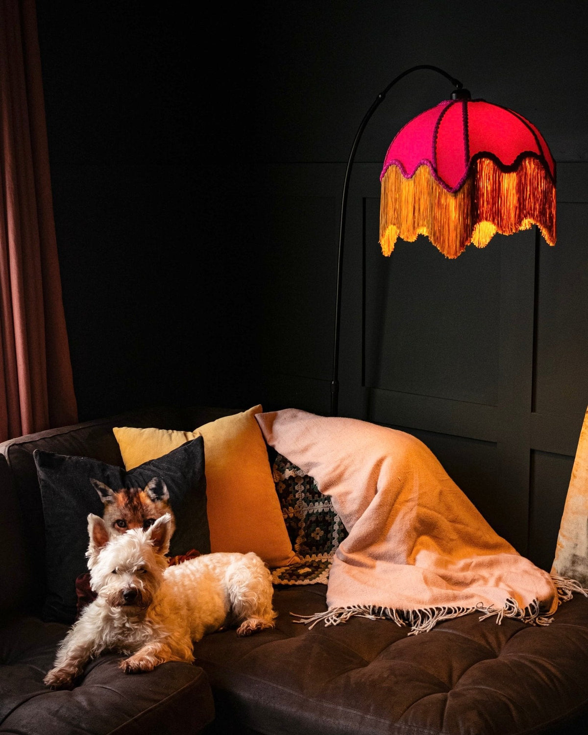 Pink and gold fringed shade on an arc floor lamp in a dark room with a westie