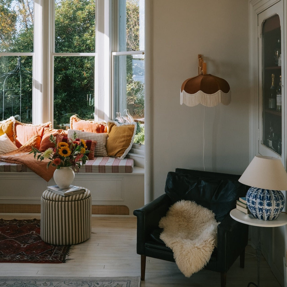 A bright sitting room with a window seat and black leather armchair, the Emmy brown and ivory fringed lampshade on the wall